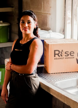 A woman standing confidently in a kitchen beside a Rise Against Hunger meal box.